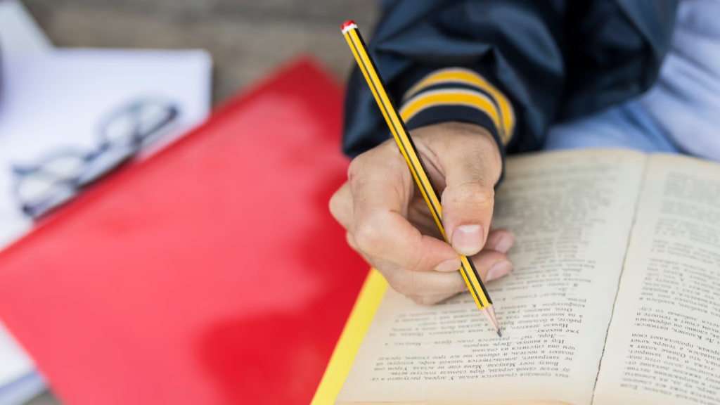 Image of a young man diligently studying a book for TOEFL test preparation, using a pen to mark down important information.
