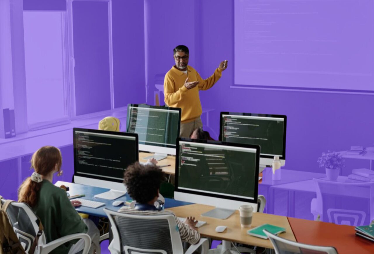 Instructor teaching programming concepts to students in a computer science classroom with multiple monitors displaying coding projects and software development exercises.